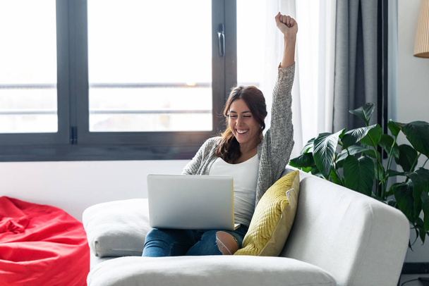 Pretty young woman celebrating something while using her laptop on sofa at home. - Foto, imagen