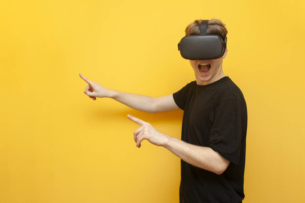 τύπος gamer σε Vr γυαλιά σε κίτρινο φόντο κατέχει μια συσκευή και δείχνει τα δάχτυλά του σε ένα μέρος για κείμενο, ένας άνθρωπος με ασύρματα gadgets για εικονική πραγματικότητα - Φωτογραφία, εικόνα