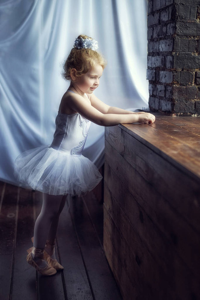 Портрет маленькой 4-летней блондинки кудрявой балерины, стоящей у окна в темной комнате в балетном платье белой пачке и пуантах. Отдых в балетной студии. Пауза в тренировке. Танцы тяжелая работа.
 - Фото, изображение