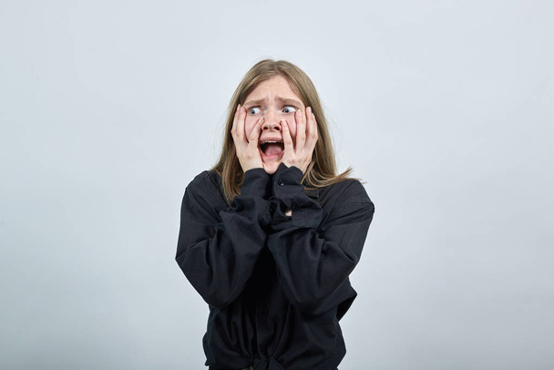 Scared young woman in fashion black shirt, keeping hands on cheeks - Photo, Image