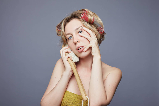 Positive blue eyed woman pretends speaking on mobile phone, holds wooden brush near ear, dressed in knitted bra, wears rollers on head, isolated on grey studio wall with free space for your promotion. - Photo, Image