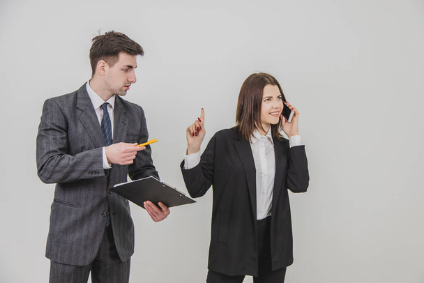 Handsome busy businesswoman is talking on the phone, pointing his finger up for her secretary to wait. Male secretary standing with clipboard, waiting for bosss signature. - Foto, imagen