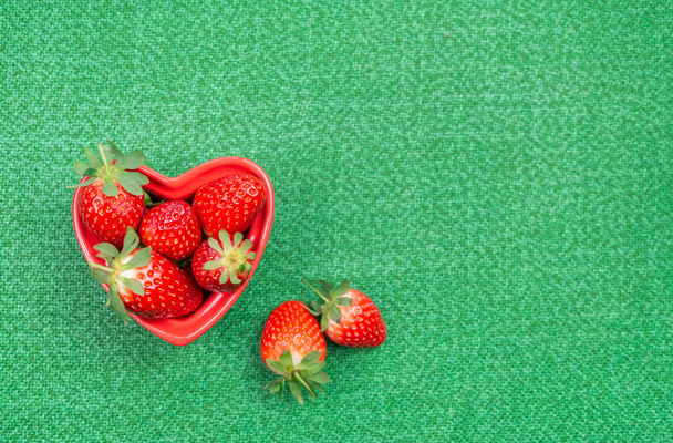 Red heart-shaped bowl with red fresh strawberries on green tablecloth. Top view, space for text - 写真・画像