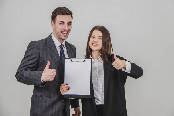 Successful business couple standing, holding together a clipboard with empty clear sheet of paper for text, giving thumbs up, smiling. - Фото, зображення