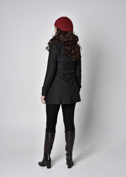 full length portrait of a pretty brunette girl wearing a black coat, leather boots and red beanie. Standing pose  on a grey studio background. - Photo, Image