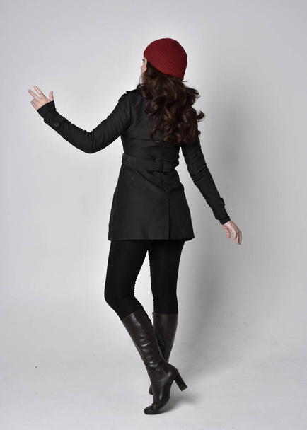 full length portrait of a pretty brunette girl wearing a black coat, leather boots and red beanie. Standing pose  on a grey studio background. - Foto, Imagem