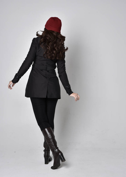 full length portrait of a pretty brunette girl wearing a black coat, leather boots and red beanie. Standing pose  on a grey studio background. - Foto, Imagen