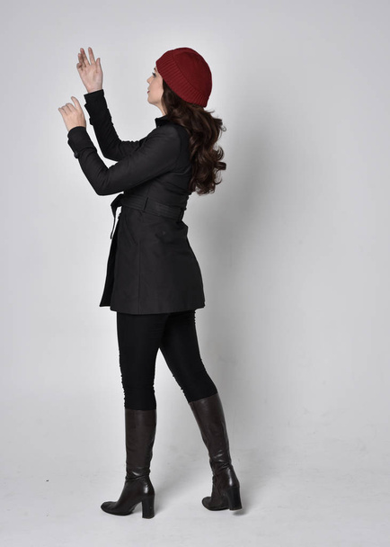 portrét krásné brunetky v černém kabátě, kožených botách a červené čepici. Stojící póza na šedém pozadí studia. - Fotografie, Obrázek
