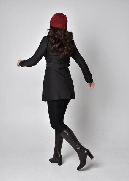 retrato de cuerpo entero de una guapa morena con un abrigo negro, botas de cuero y gorro rojo. Posar de pie sobre un fondo gris de estudio
. - Foto, imagen