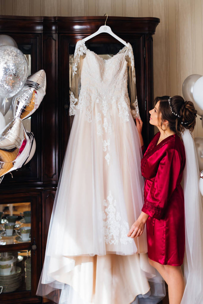 брюнетка наречена тримає білу весільну сукню на вішалці. кімната відьма
 - Фото, зображення