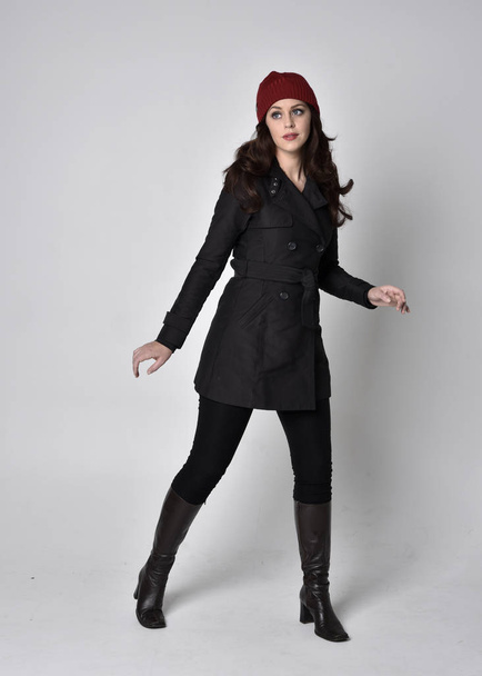 full length portrait of a pretty brunette girl wearing a black coat, leather boots and red beanie. Standing pose  on a grey studio background. - Fotografie, Obrázek