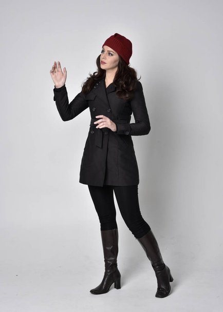 full length portrait of a pretty brunette girl wearing a black coat, leather boots and red beanie. Standing pose  on a grey studio background. - Фото, изображение