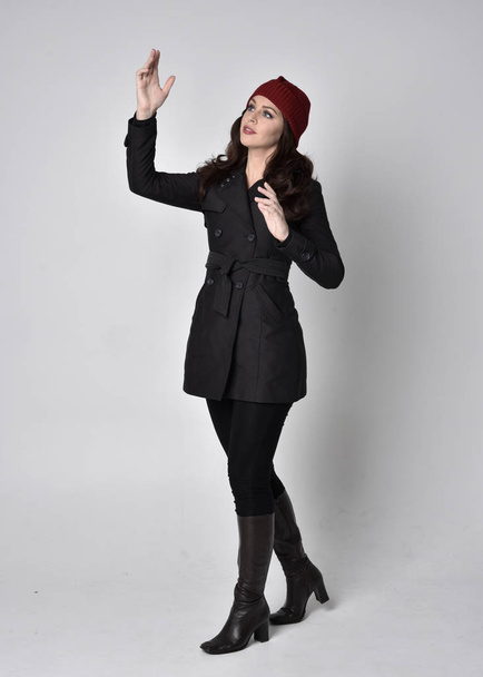 ritratto a figura intera di una bella ragazza bruna che indossa un cappotto nero, stivali di pelle e berretto rosso. In piedi posa su uno sfondo grigio studio
. - Foto, immagini