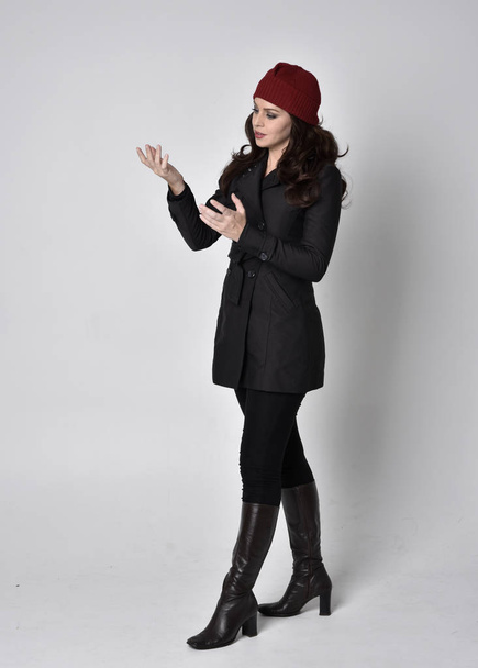 volledig portret van een mooi brunette meisje met een zwarte jas, leren laarzen en rode muts. Staande houding op een grijze studio achtergrond. - Foto, afbeelding