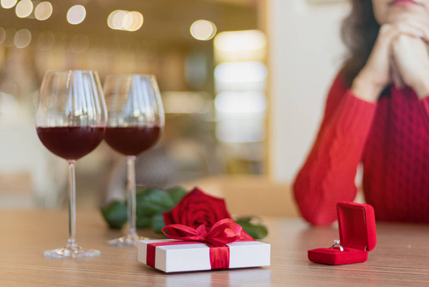 Beyaz hediye kutusu, iki şarap kadehi, masada duran bir gül ve bir yüzük. Kırmızı bir kazak giyen genç bir kadın gülümsüyor. Kırmızı kurdeleli bir hediye. Sevgililer Günü konsepti. - Fotoğraf, Görsel