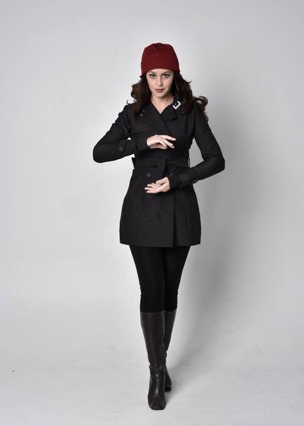 full length portrait of a pretty brunette girl wearing a black coat, leather boots and red beanie. Standing pose  on a grey studio background. - 写真・画像