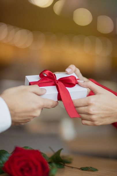 A young woman takes a present giftbox with red ribbon from her boyfriend. Warm and lovely background of a restaurant. Two glasses of wine and a rose on the cafe's table. Valentine's day concept. - Фото, изображение