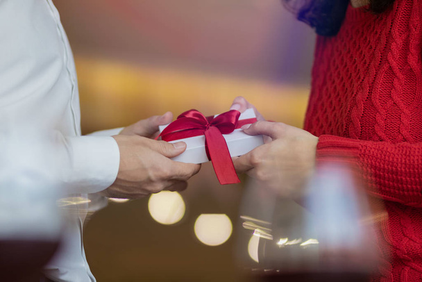 A man passes a white present giftbox with a red ribbon to his girlfriend's hands. Classic look of a couple, red sweater and white shirt. Valentine's day and winter holidays concept. - Photo, image