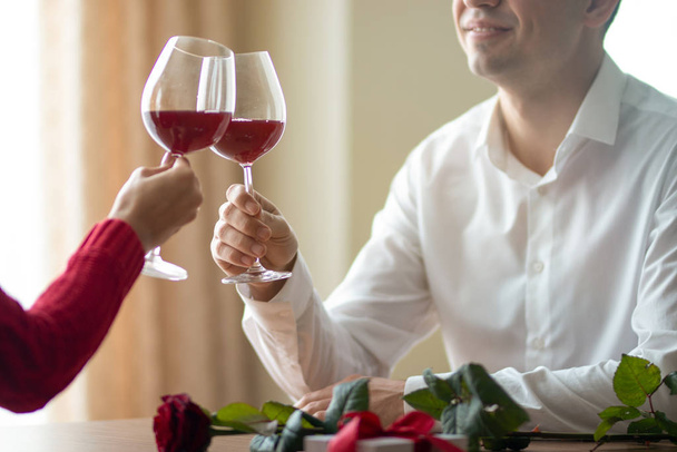 Veselý mladý pár přecházející přes skleničky v restauraci. Muž a jeho přítelkyně popíjejí víno v kavárně. Rudá růže na stole. Valentýnský koncept. - Fotografie, Obrázek
