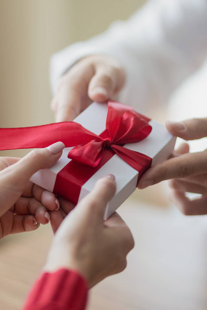 A small present is being passed by a couple from one to another. White little giftbox with a red ribbon in the hands. Valentine's day and winter holidays concept. - 写真・画像