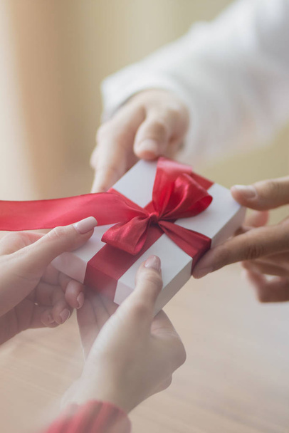 A small present is being passed by a couple from one to another. White little giftbox with a red ribbon in the hands. Valentine's day and winter holidays concept. - Foto, immagini