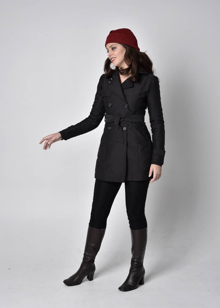 full length portrait of a pretty brunette girl wearing a black coat, leather boots and red beanie. Standing pose  on a grey studio background. - Fotografie, Obrázek