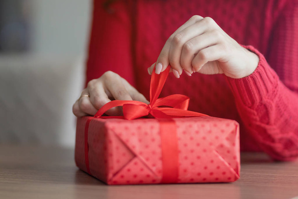A young woman opens a present at a cafe. Red giftbox with a ribbon is being untied on a restaurant's table. Valentine's day and winter holidays concept. - 写真・画像