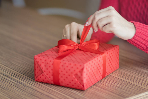 Μια νεαρή γυναίκα ανοίγει ένα δώρο σε ένα καφέ. Το κόκκινο κουτί δώρου με την κορδέλα λύνεται στο τραπέζι ενός εστιατορίου. Ημέρα του Αγίου Βαλεντίνου και χειμερινές διακοπές έννοια. - Φωτογραφία, εικόνα
