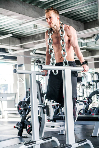 Αθλητής με μια τεράστια μεταλλική αλυσίδα στο λαιμό του. Push-ups σχετικά με την άνιση μπαρ στο γυμναστήριο - Φωτογραφία, εικόνα