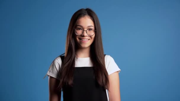 Pretty young asian girl with long hair standing on blue studio background. Cute portrait of woman in transparent round glasses and black overalls. - Felvétel, videó