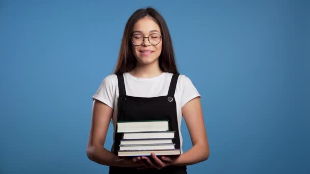 Asian student girl on blue background in the studio holds stack of university books from library. Woman smiles, she is happy to graduate. - Video