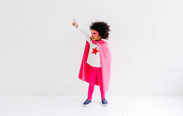 Πορτρέτο της Αφρικής Αμερικής κοριτσάκι παίζει superhero στο φόντο λευκό τοίχο. Έννοια δύναμης κοριτσιών. - Φωτογραφία, εικόνα