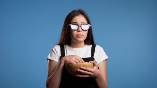 Young asian woman in 3d glasses watching horror movie and eating popcorn on blue studio background. Frightened girl because of unexpectedness plot in film. - Imágenes, Vídeo