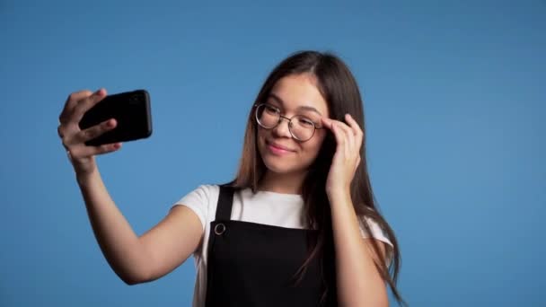 Χαμογελώντας ευτυχισμένη Ασιάτισσα με γυαλιά κάνοντας selfie στο smartphone πάνω από το μπλε φόντο. Τεχνολογία, κινητή συσκευή, έννοια των κοινωνικών δικτύων. - Πλάνα, βίντεο