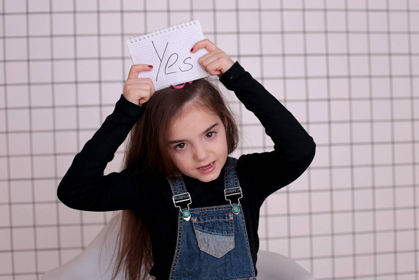 mladá hravá osmiletá dívka s dlouhými vlasy v černém roláku a džínovými šaty s nápisem "Ano" - Fotografie, Obrázek