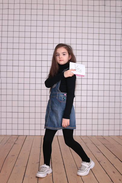 mladá hravá osmiletá dívka s dlouhými vlasy v černém roláku a džínovými šaty s nápisem "Ano" - Fotografie, Obrázek