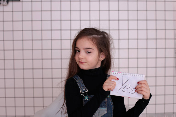 jong soort speels meisje van acht jaar met lang haar in een zwarte coltrui en denim sundress met het teken "Ja" - Foto, afbeelding