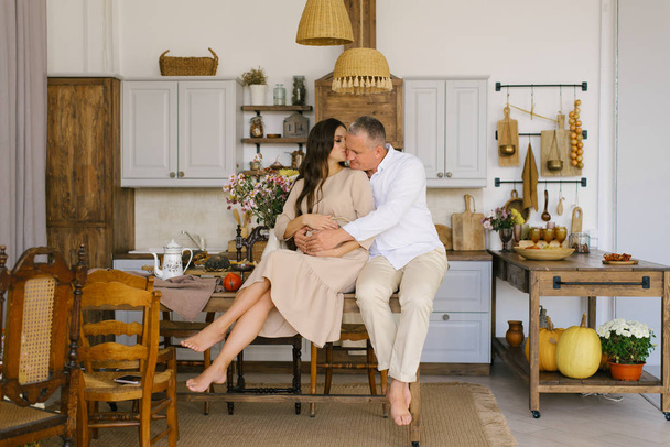 супружеская пара обнимается, сидя на столе на кухне. Муж обнимает беременную жену и кладет руки на её большой живот.
. - Фото, изображение