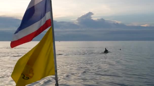 Akşamları Tayland bayraklarının yanında yüzen tekne. Tayland ve Tayland Kralı 'nın bayrakları, bulutlu gün batımına karşı dalgalı denizde yüzen isimsiz bir insan silueti.. - Video, Çekim