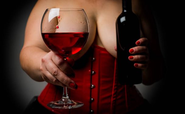 Plus size woman with big beautiful breast holding a glass with wine and bottle of wine. Sexy hot woman with big boobs wearing red corset in Victorian style. Sweet wine and woman concept. - Foto, afbeelding