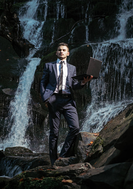Привлекательный человек в костюме держит ноутбук на заднем плане водопада. Красивый молодой бизнесмен стоит под водопадом со своим инновационным устройством с водонепроницаемой защитой IP68
. - Фото, изображение