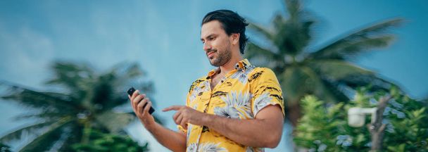 Un hombre contento está de pie con un teléfono en sus manos sobre un fondo de palmeras
 - Foto, imagen