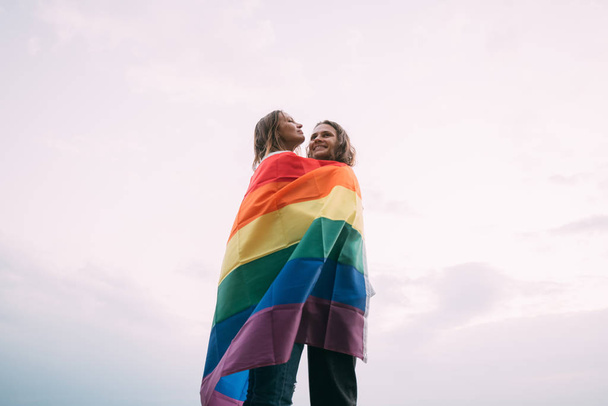 Dos mujeres con bandera de arco iris en la playa contra el cielo
. - Foto, imagen