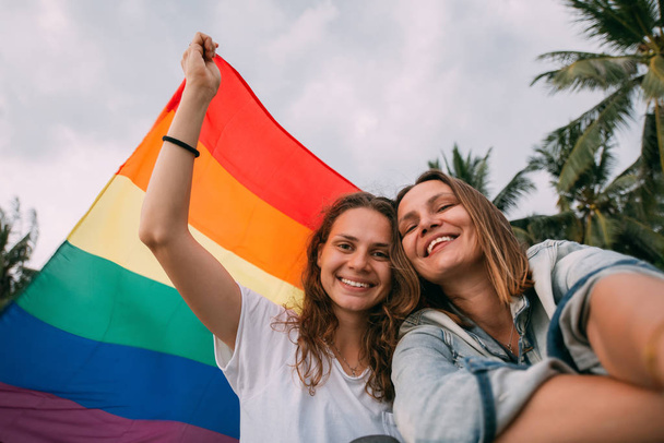 Two women with rainbow flag on the beach on a background of palm trees - Fotoğraf, Görsel