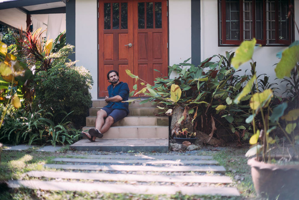 Een man zit op de veranda van een huis in een tropische tuin - Foto, afbeelding