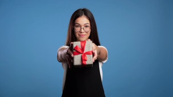 Pretty brunette asian girl in glasses gives gift and hands it to the camera. She is happy, smiling. Girl on blue background. Positive holiday footage - Imágenes, Vídeo