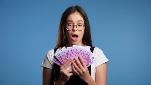 Ικανοποιημένο χαρούμενο ενθουσιασμένο κορίτσι από την Ασία που δείχνει χρήματα - χαρτονομίσματα του ευρώ στον μπλε τοίχο. Σύμβολο επιτυχίας, κέρδους, νίκης. - Πλάνα, βίντεο