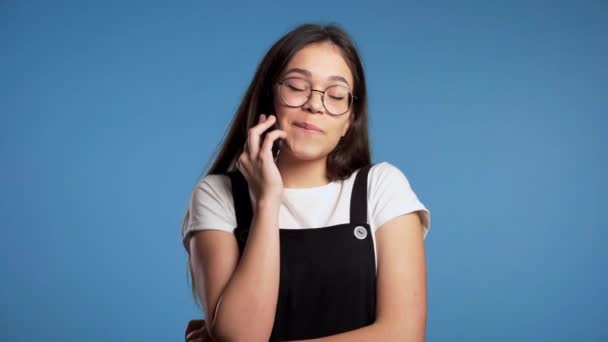 Pretty asian girl with long hair speaks on phone and laughs infectiously from interlocutors jokes. Trendy outfit. Blue studio background - Filmagem, Vídeo