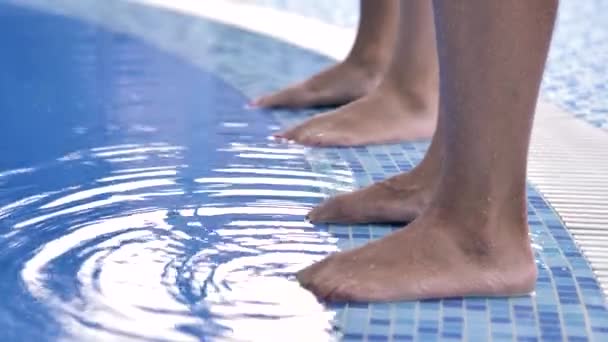 Two pairs of female legs stand on the side of the pool with blue clear water and move their fingers. - Video