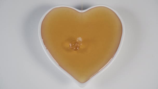Filling with yellow bee honey a white plate in the shape of a heart on a white kitchen table. A thin stream flows slowly. - Imágenes, Vídeo